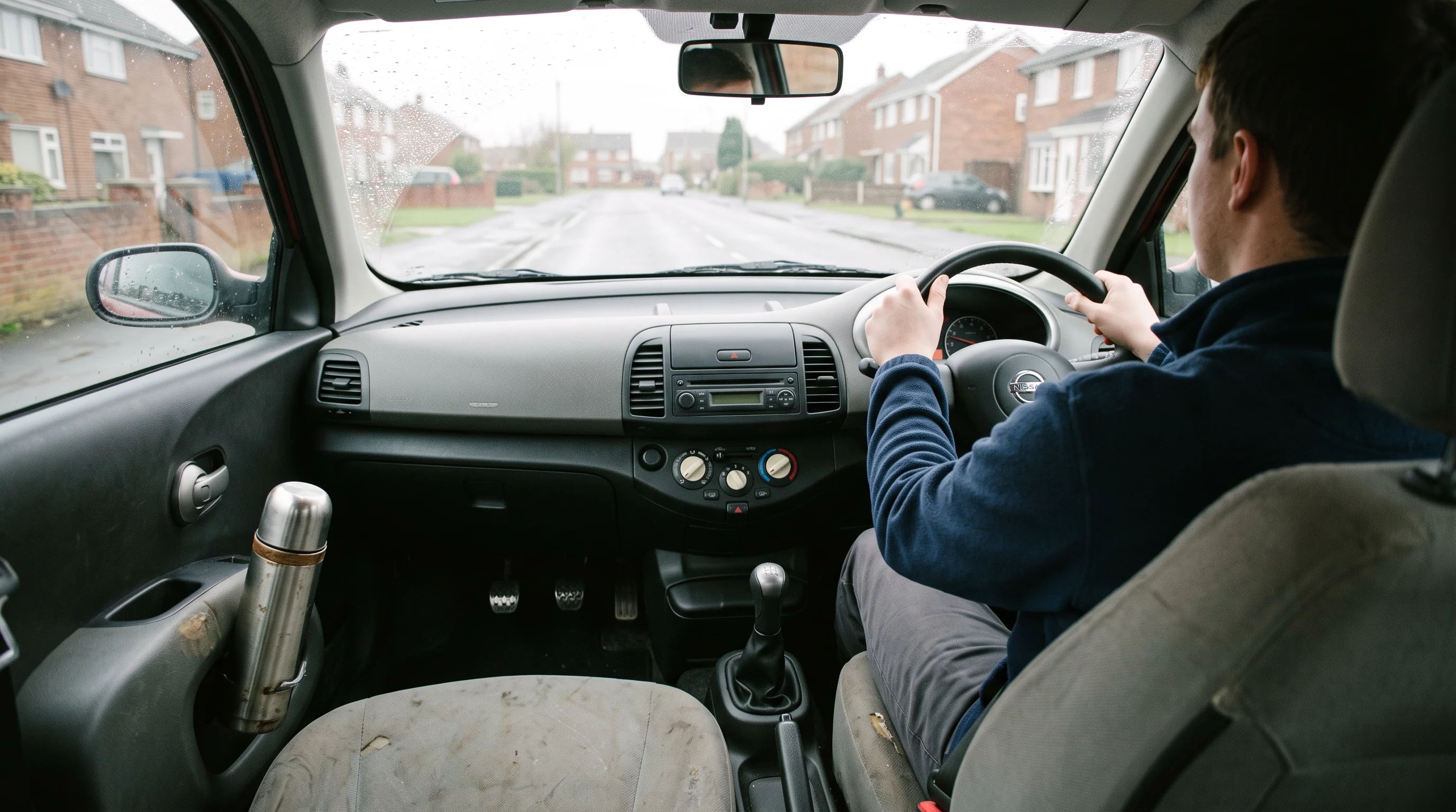 A learner's hands resting at ten-to-two on the steering wheel of a tuition car, with dual-control pedals in the footwell and a flask in the door pocket